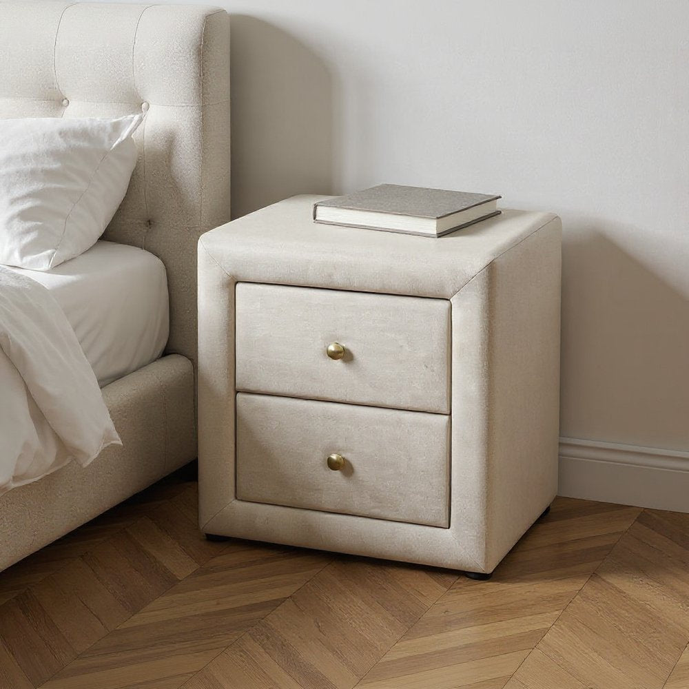 Beige wood two-drawer nightstand with brass knobs, a book on top beside a tufted bed.