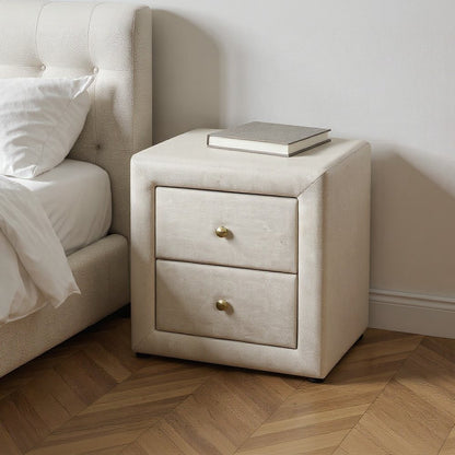Beige wood two-drawer nightstand with brass knobs, a book on top beside a tufted bed.