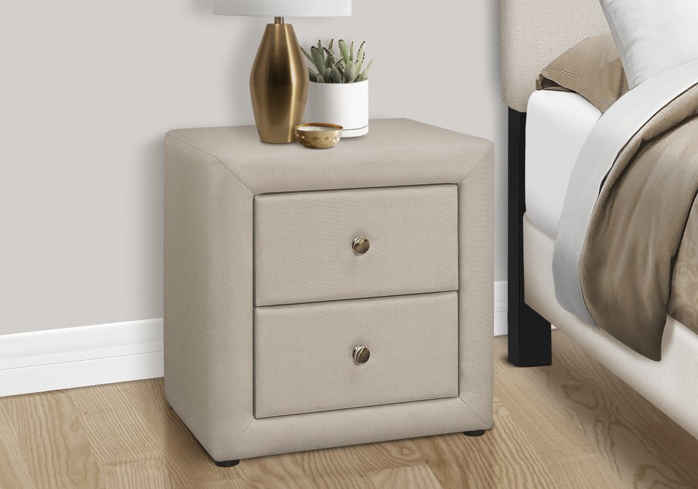 Beige wood two-drawer nightstand with round metal knobs, in a bedroom beside a bed with a gold vase and plant.