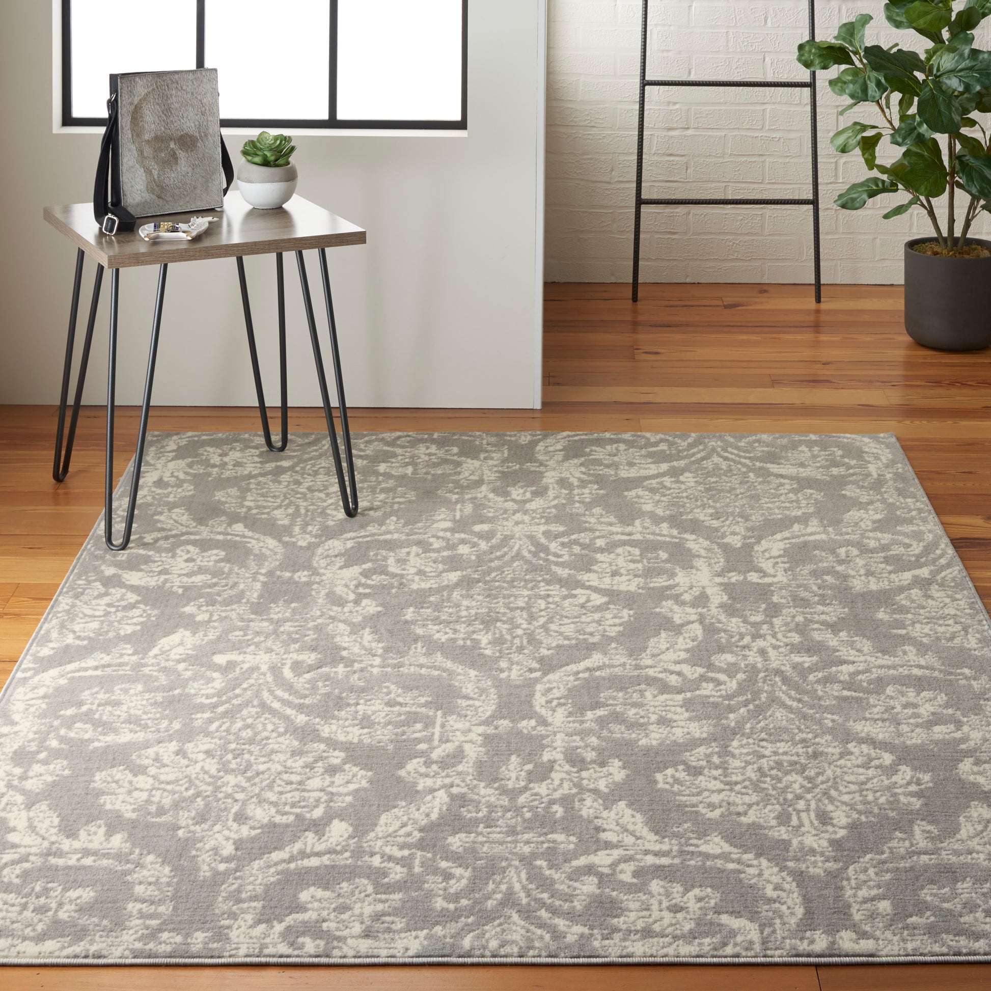 Gray ornate patterned area rug on warm wood floor with a small wood desk, black metal legs, and potted plants.