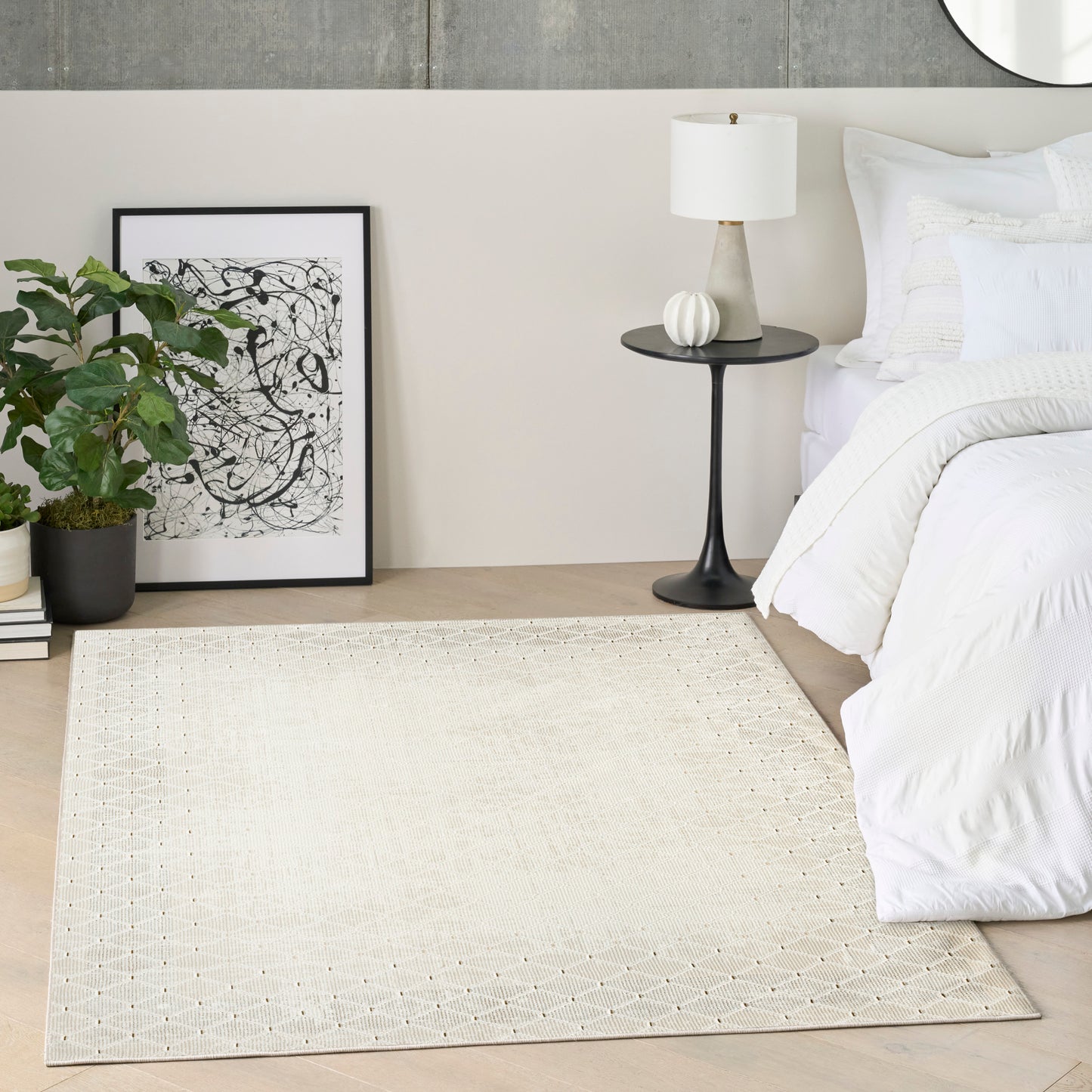 Neutral bedroom with beige dotted rug, white bedding, black round nightstand & white lamp, abstract art & a potted plant.