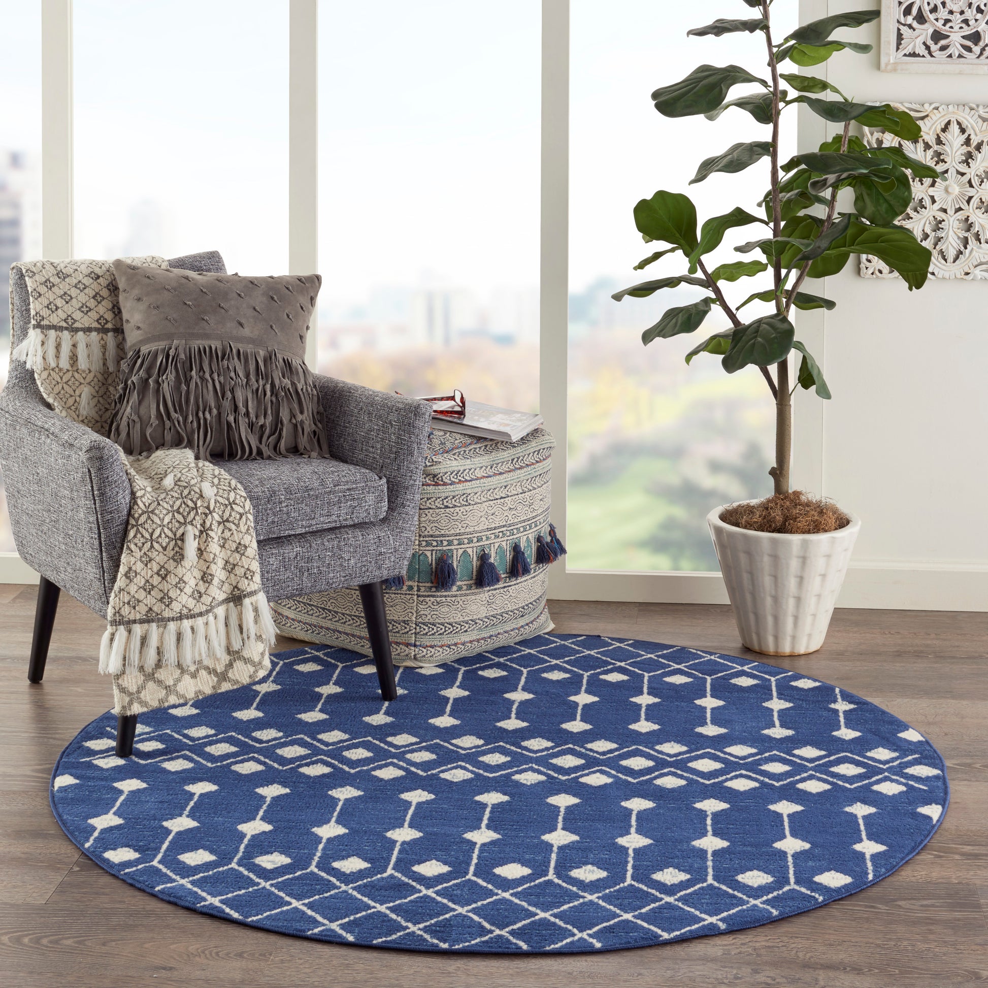 Blue round rug with white geometric pattern under a gray armchair; fringed throw and a tall potted plant nearby.