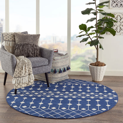 Blue round rug with white geometric pattern under a gray armchair; fringed throw and a tall potted plant nearby.