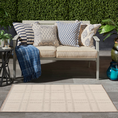 Outdoor beige cushioned sofa with patterned pillows, blue throw, beige rug, garden backdrop.