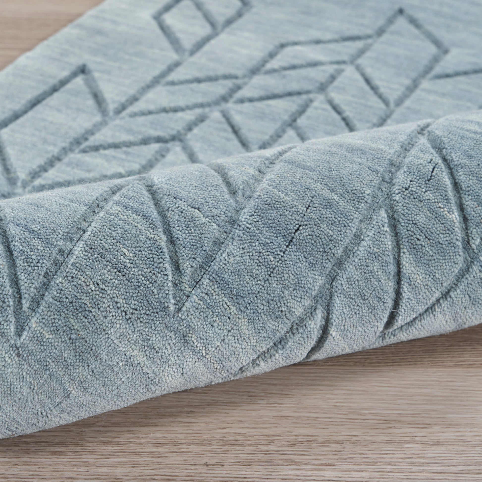 Blue-gray geometric area rug with raised linear pattern, rolled corner on wood floor.