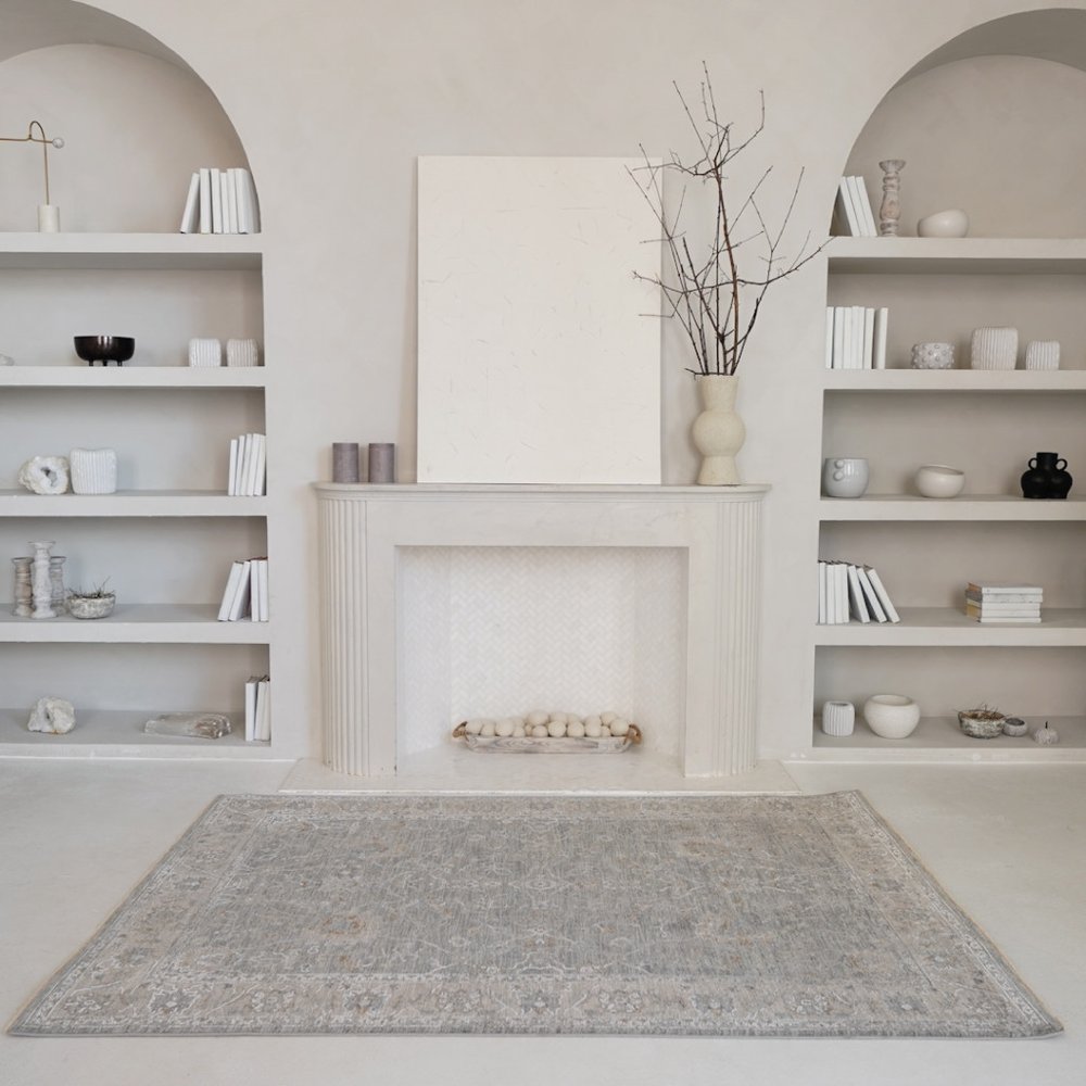 Minimalist living room with white mantel and built-in shelves, bare branches in a vase, large white canvas, and a faded gray patterned rug.