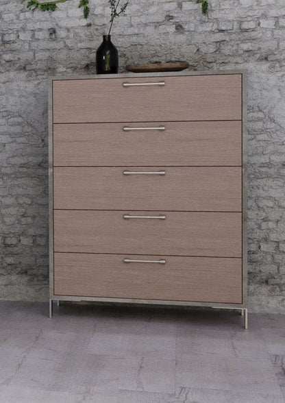 Light wood five-drawer chest with silver handles on slim metal legs, brick wall backdrop, decorative vase on top.