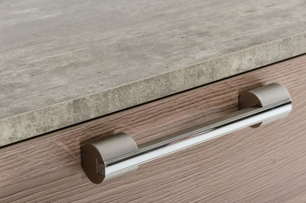 Modern chrome cylindrical pull handle on a light wood cabinet drawer beneath a gray stone countertop.