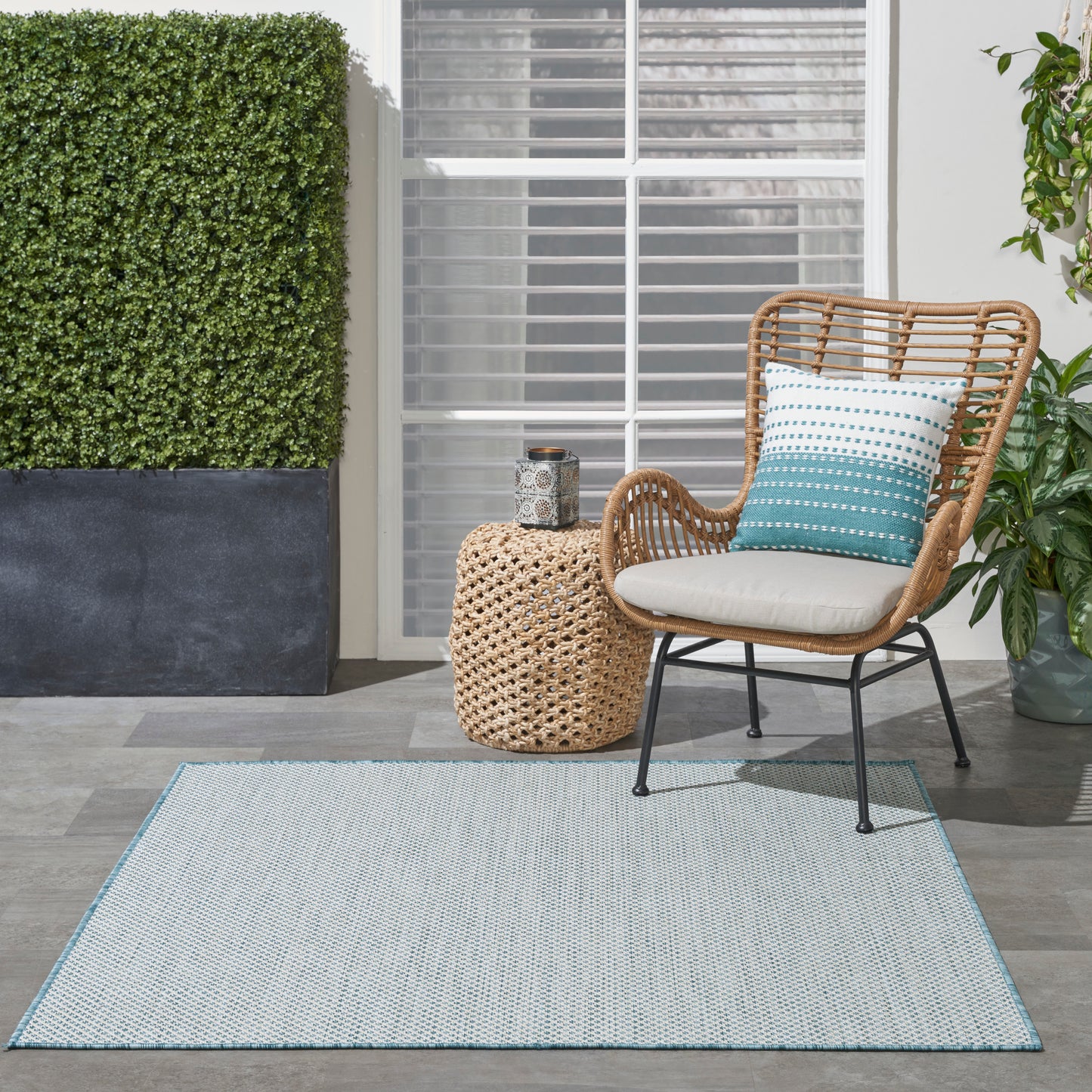 Outdoor wicker chair with white cushion, teal striped pillow, round woven side table, light blue rug, and greenery.