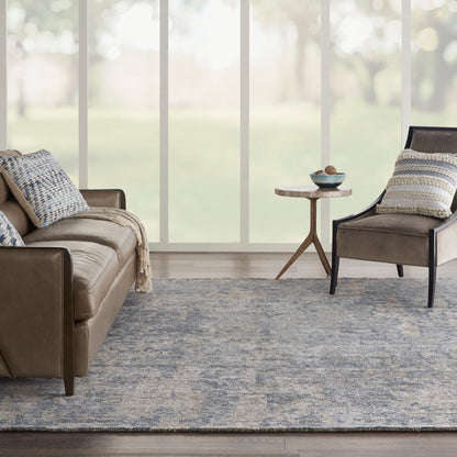 Blue-gray patterned rug in a living room with a brown leather sofa and armchair and a round wooden side table by large windows.