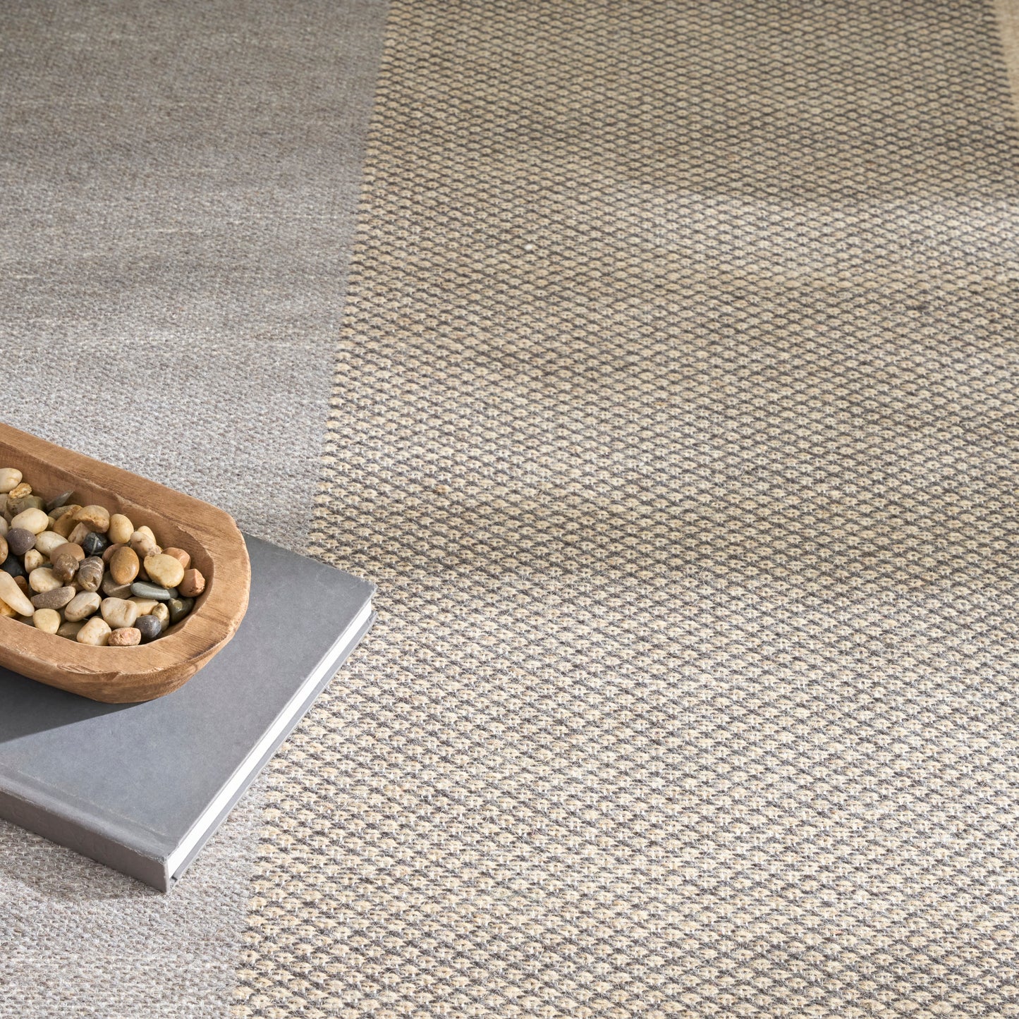 Two-tone woven rug: light gray left panel and beige textured patterned right; wooden tray with pebbles atop a gray book.