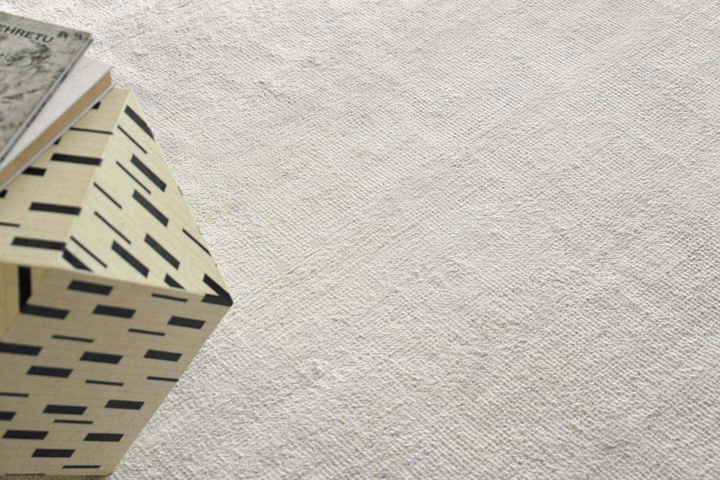 Beige plush indoor hand-knotted rug with a light textured surface and a black rectangular dash pattern, angled with a small book resting on it, 2' x 3'.