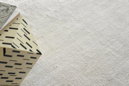 Beige plush indoor hand-knotted rug with a light textured surface and a black rectangular dash pattern, angled with a small book resting on it, 2' x 3'.
