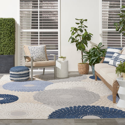 Neutral outdoor lounge with beige sofa, armchairs, blue circular rug, potted greenery, and woven baskets.
