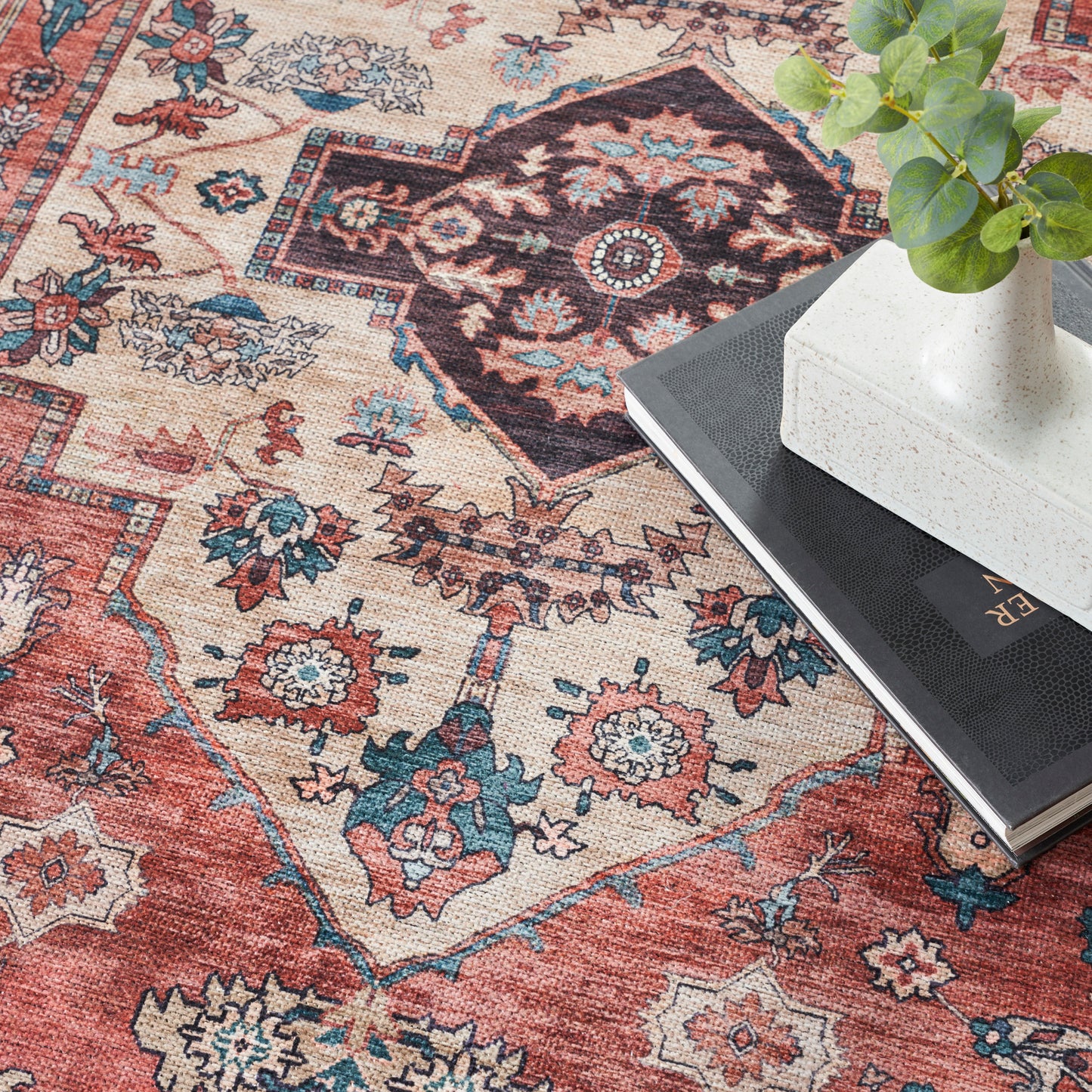 Colorful oriental rug with red, blue and beige patterns; white vase with green plant atop a black book.