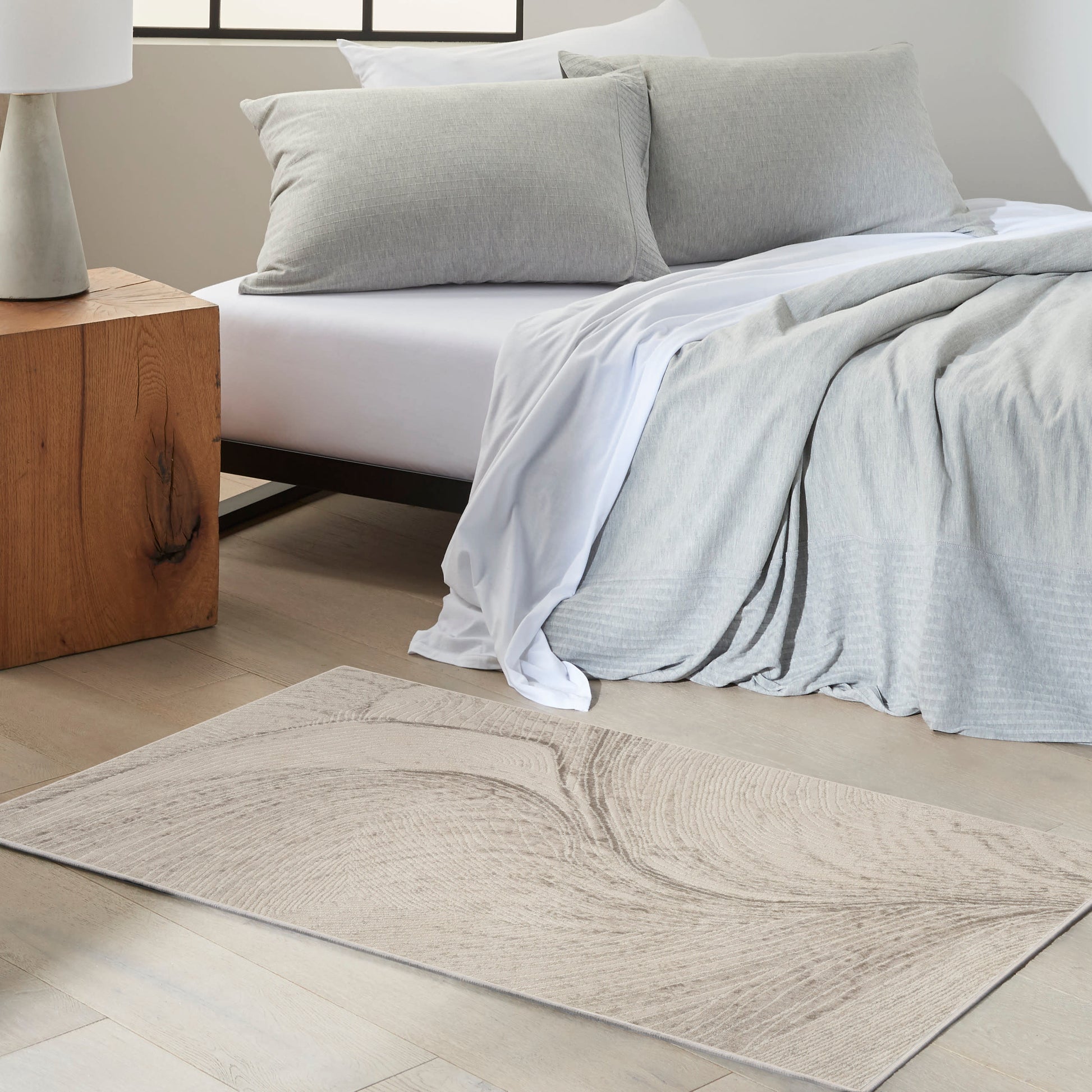 Soft gray bedding with white sheets and matching gray pillows on a bed; light wood nightstand and beige swirled rug.