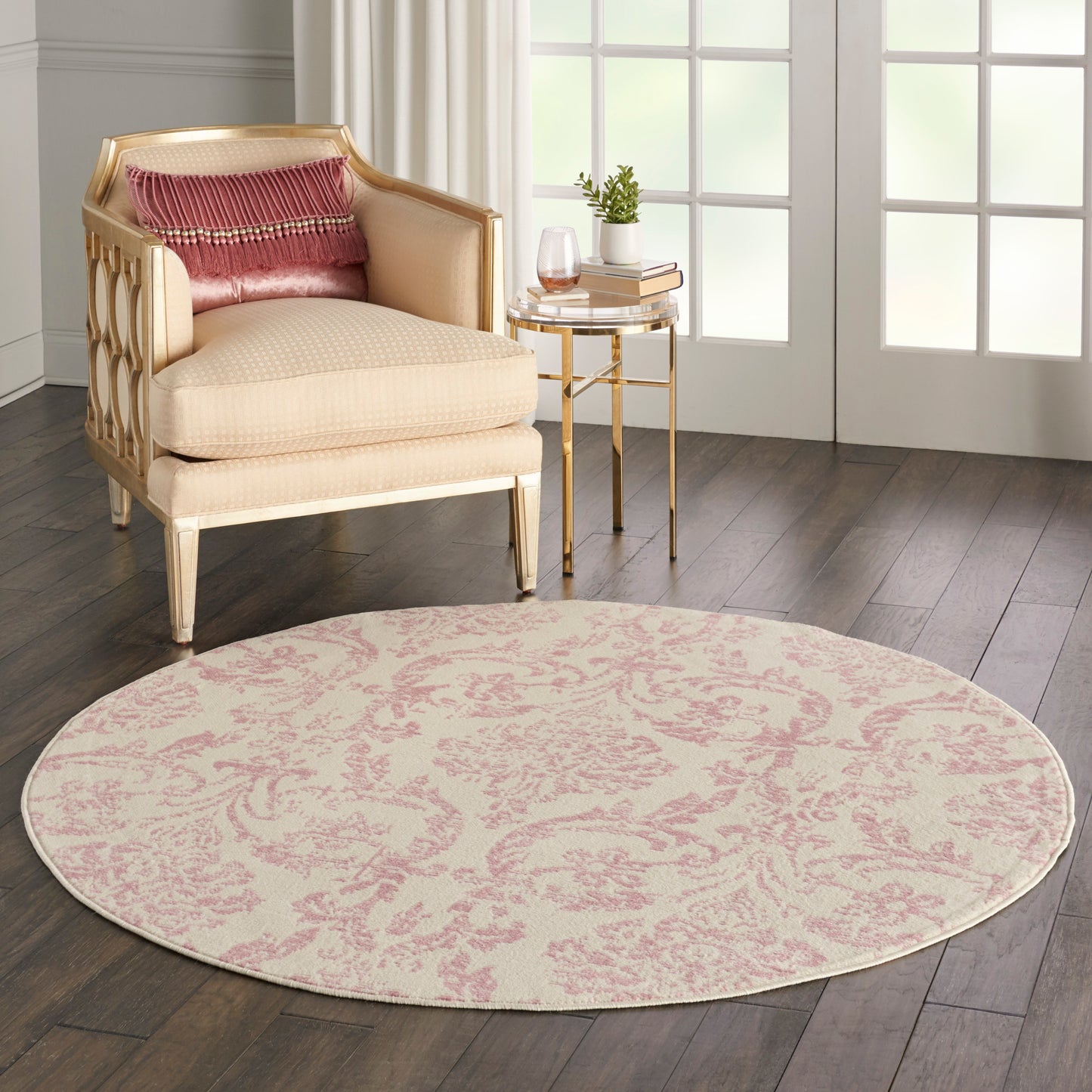 Round pink damask area rug, beige upholstered armchair with pink cushions, and gold glass-top side table.