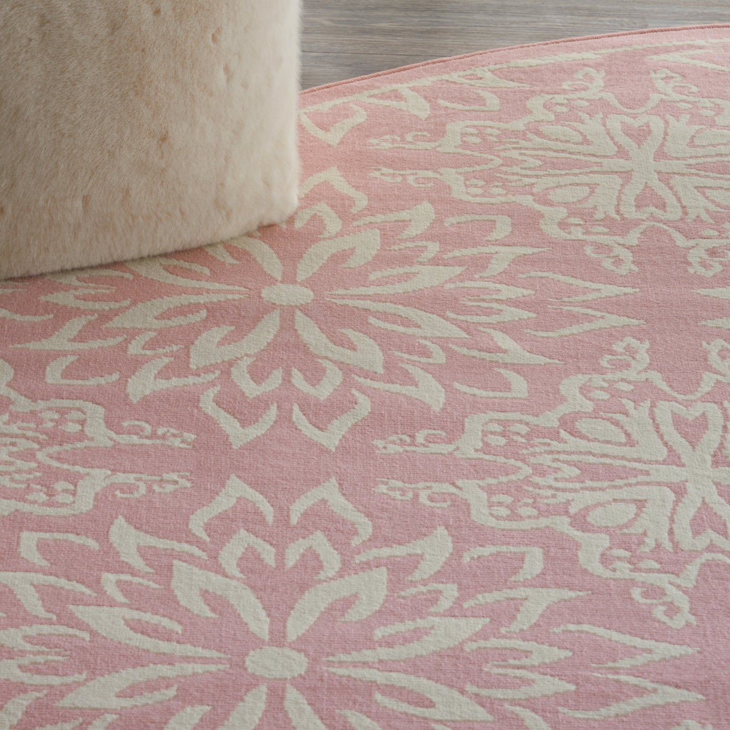Round pink rug with white mandala floral pattern; soft wool texture, beige faux fur stool at edge.
