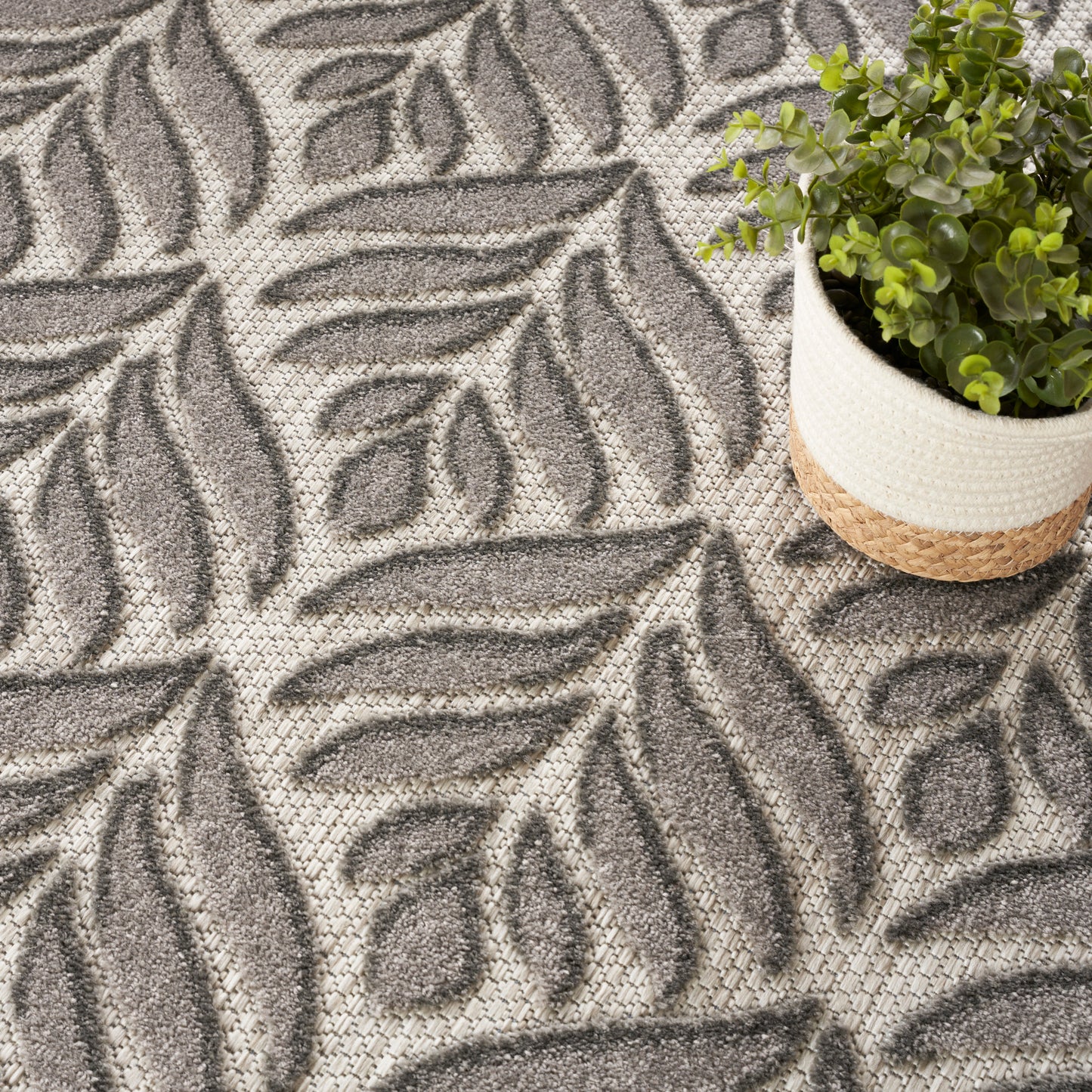 Neutral gray tropical leaf-pattern rug with beige woven background; plant in a two-tone jute basket.