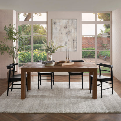 Rustic wooden dining table with black chairs on a light shag rug in a dining room; abstract wall art, greenery outside.