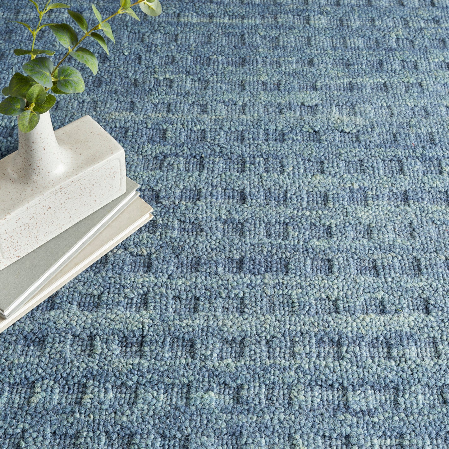 Blue boucle rug with chunky loops; white speckled vase with greenery atop neutral stacked books.