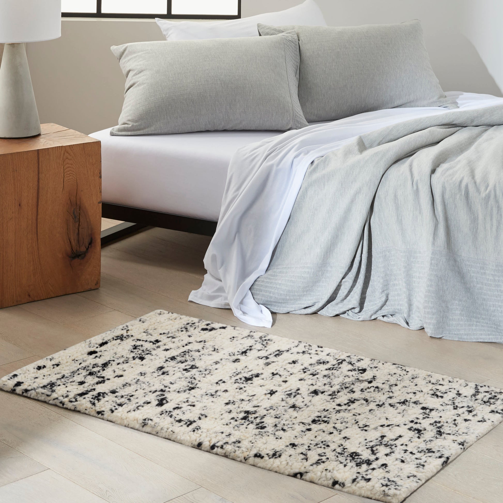 Bedroom with a light gray duvet and pillows on white bedding, a wooden side table, and a cream speckled rectangular rug.