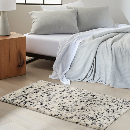 Bedroom with a light gray duvet and pillows on white bedding, a wooden side table, and a cream speckled rectangular rug.