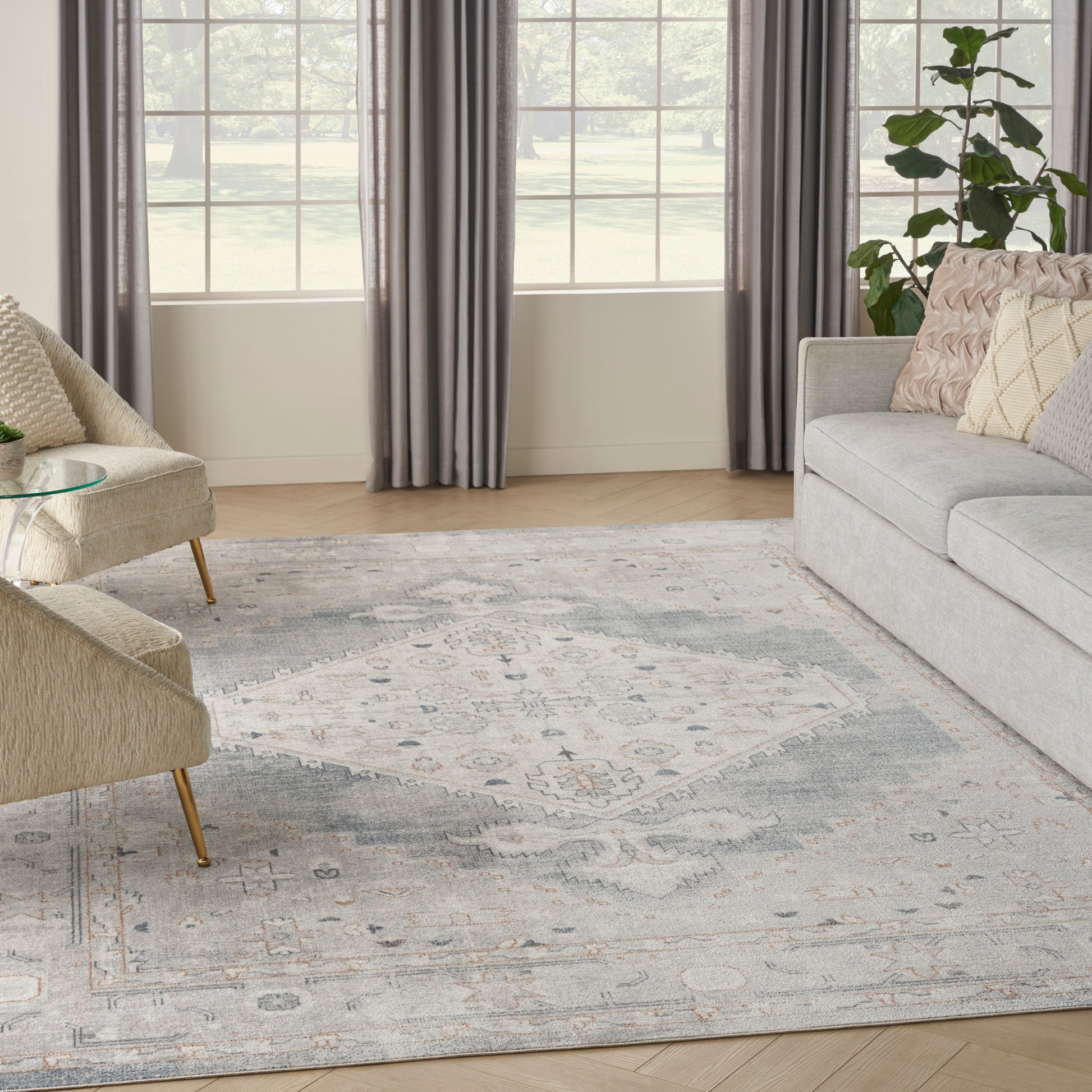 Cozy living room with light gray sofa, beige accent chairs, glass coffee table, blue-gray rug, gray curtains, and a plant.