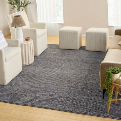 Living room with a dark gray textured area rug, cream sofas, two square ottomans, wooden side table with lamp, and greenery.