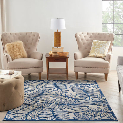 Two beige tufted armchairs with pillows flank a wooden side table and lamp; blue leaf-pattern rug on light wood floor.