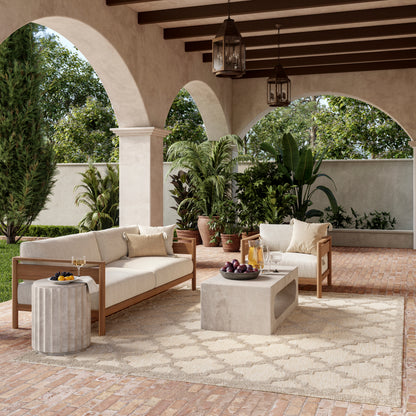 Outdoor Mediterranean patio with beige cushioned wooden sofas, concrete cube coffee table, rug, and lush potted plants.