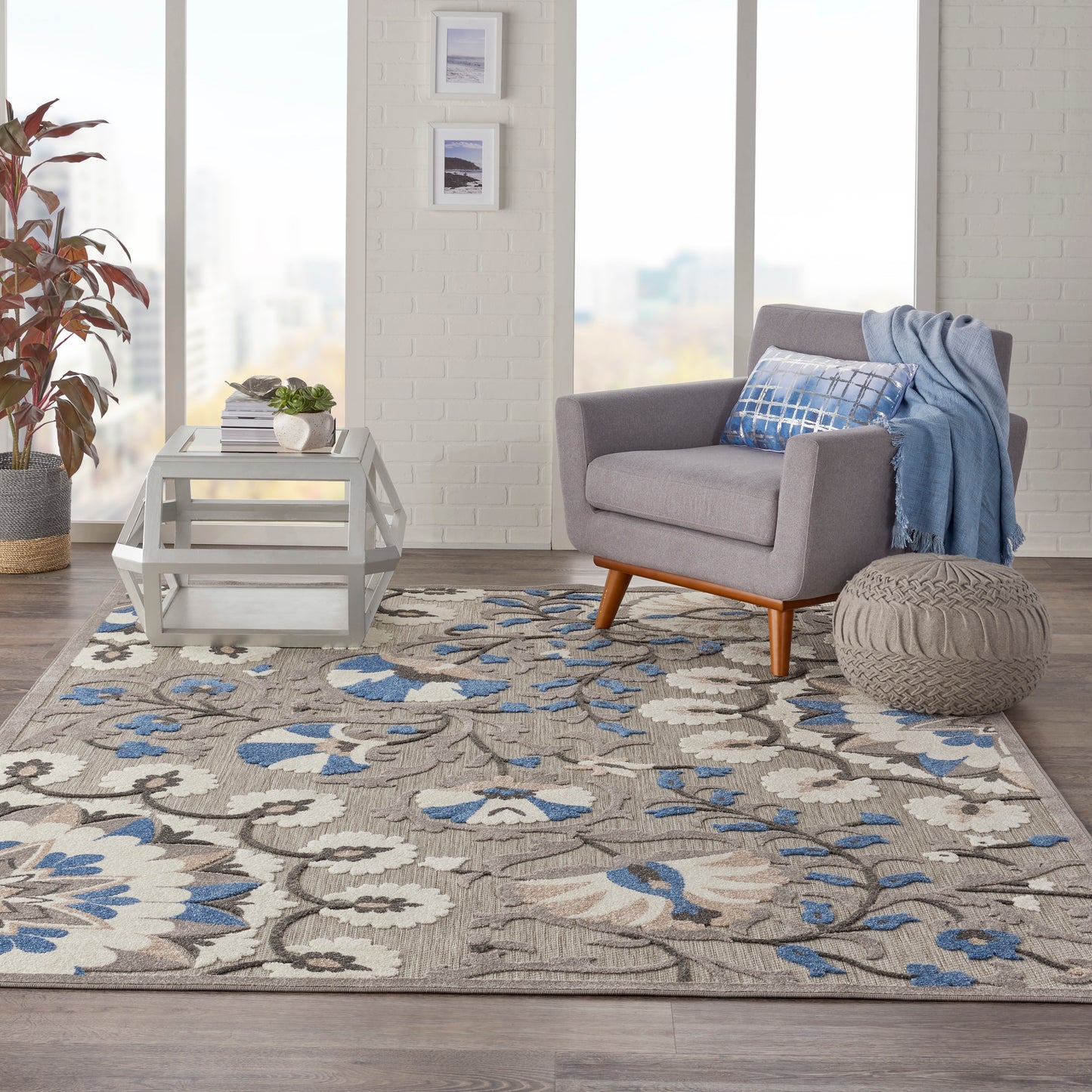 Beige-blue floral rug (8' x 11') in a living room scene with a gray mid-century chair, blue throw, white coffee table, knitted ottoman, and a plant.