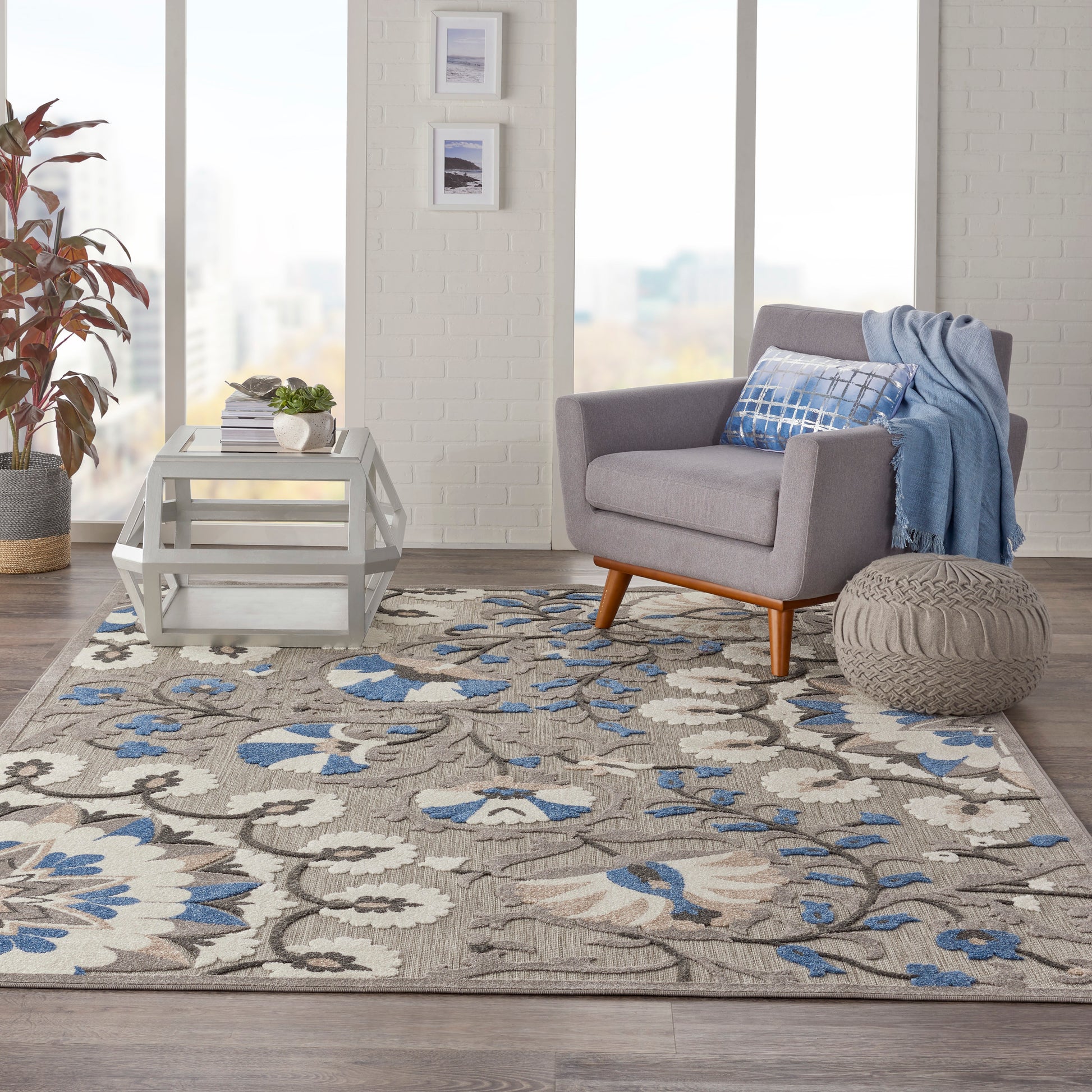 Beige-blue floral rug (8' x 11') in a living room scene with a gray mid-century chair, blue throw, white coffee table, knitted ottoman, and a plant.