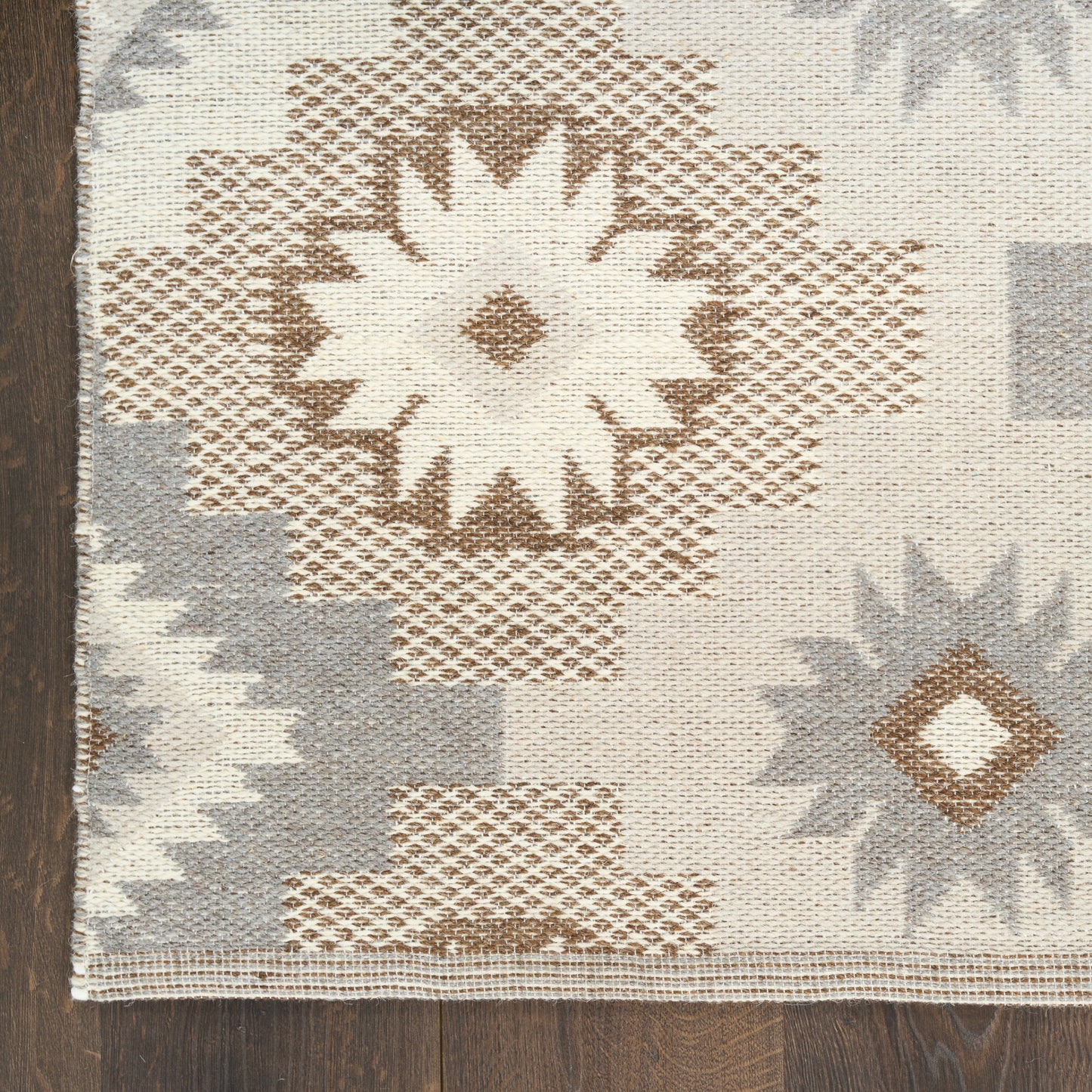 Geometric woven rug with large star motifs in beige, brown, and gray on neutral cream background, on dark wood floor.