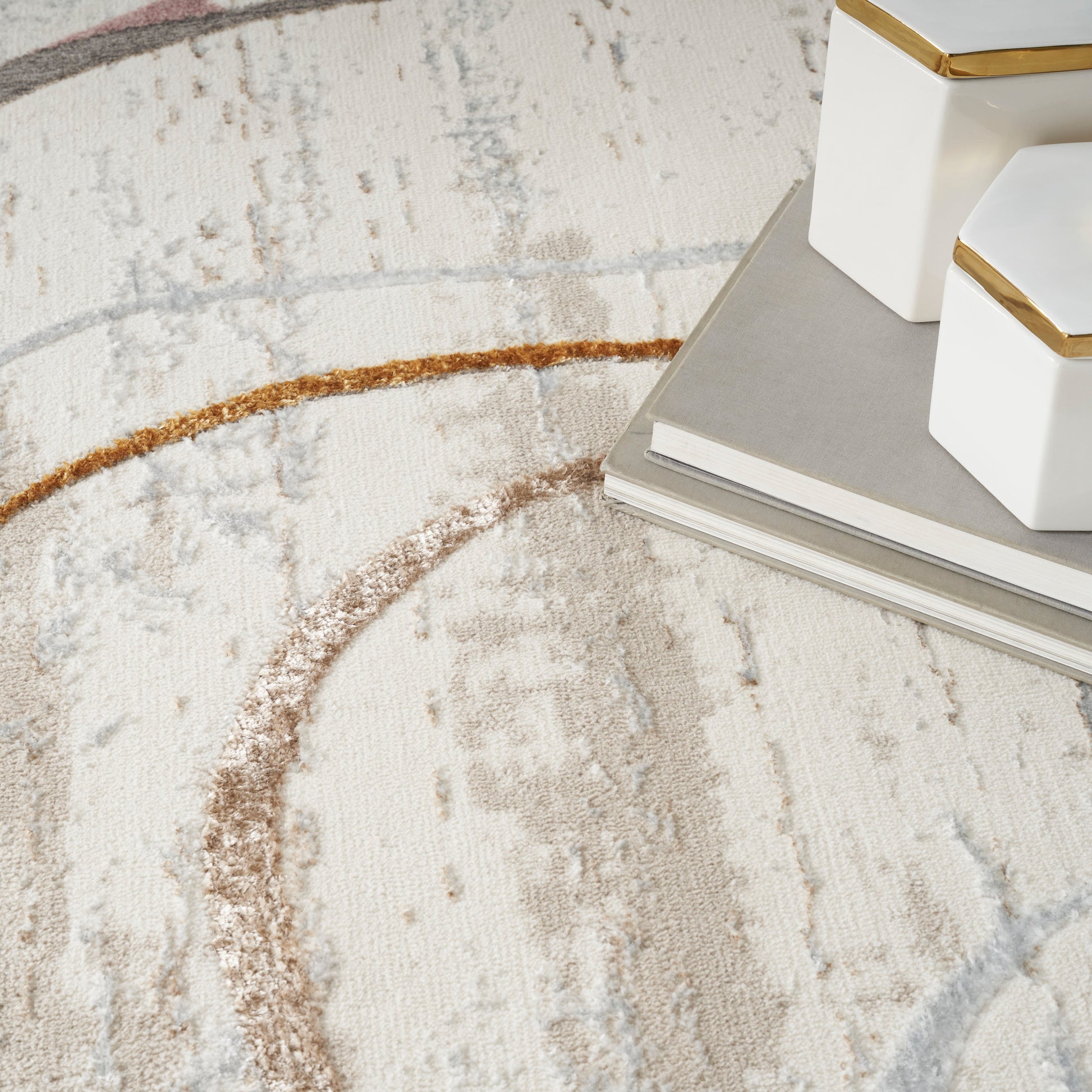 Cream rug with beige and taupe circular arc patterns; white ceramic boxes with gold trim atop gray notebooks.
