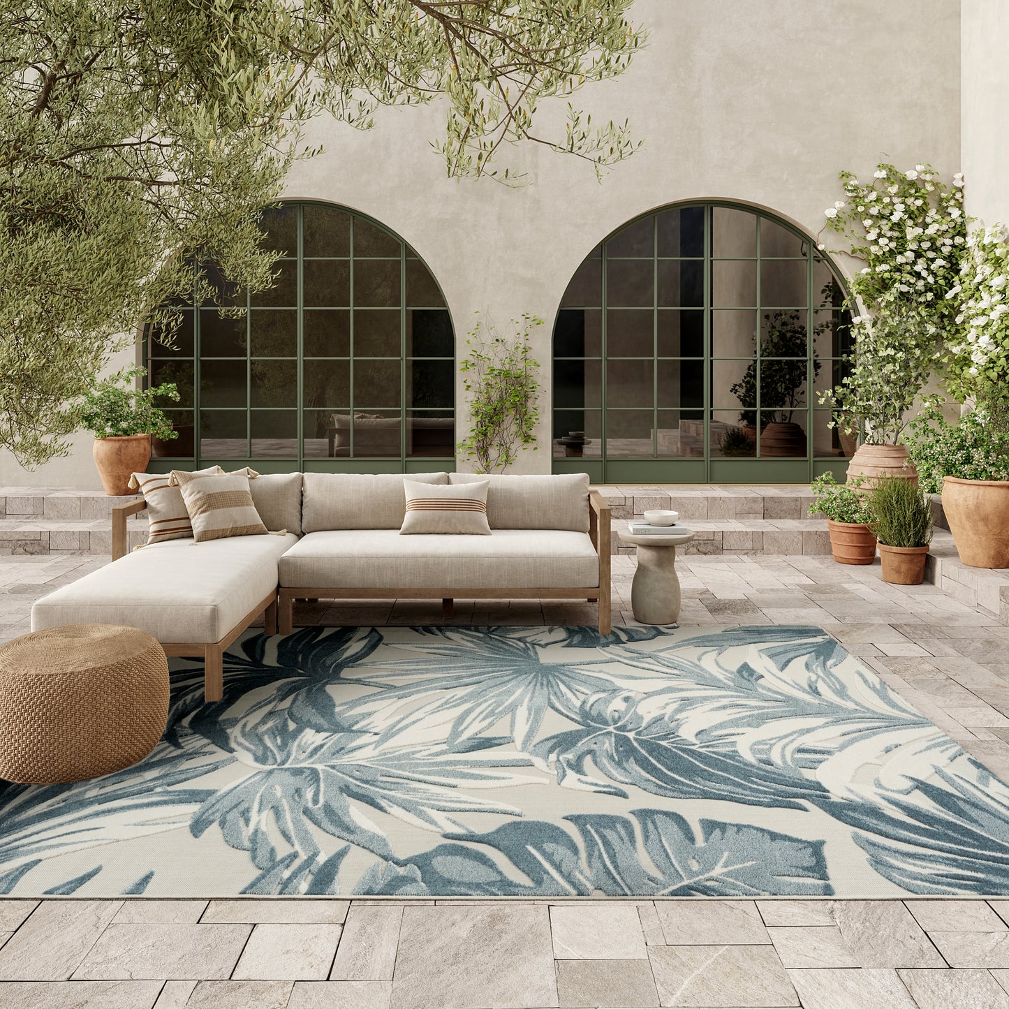 Outdoor beige sectional sofa with cushions on a blue palm-leaf rug, arched green-framed windows and potted plants.