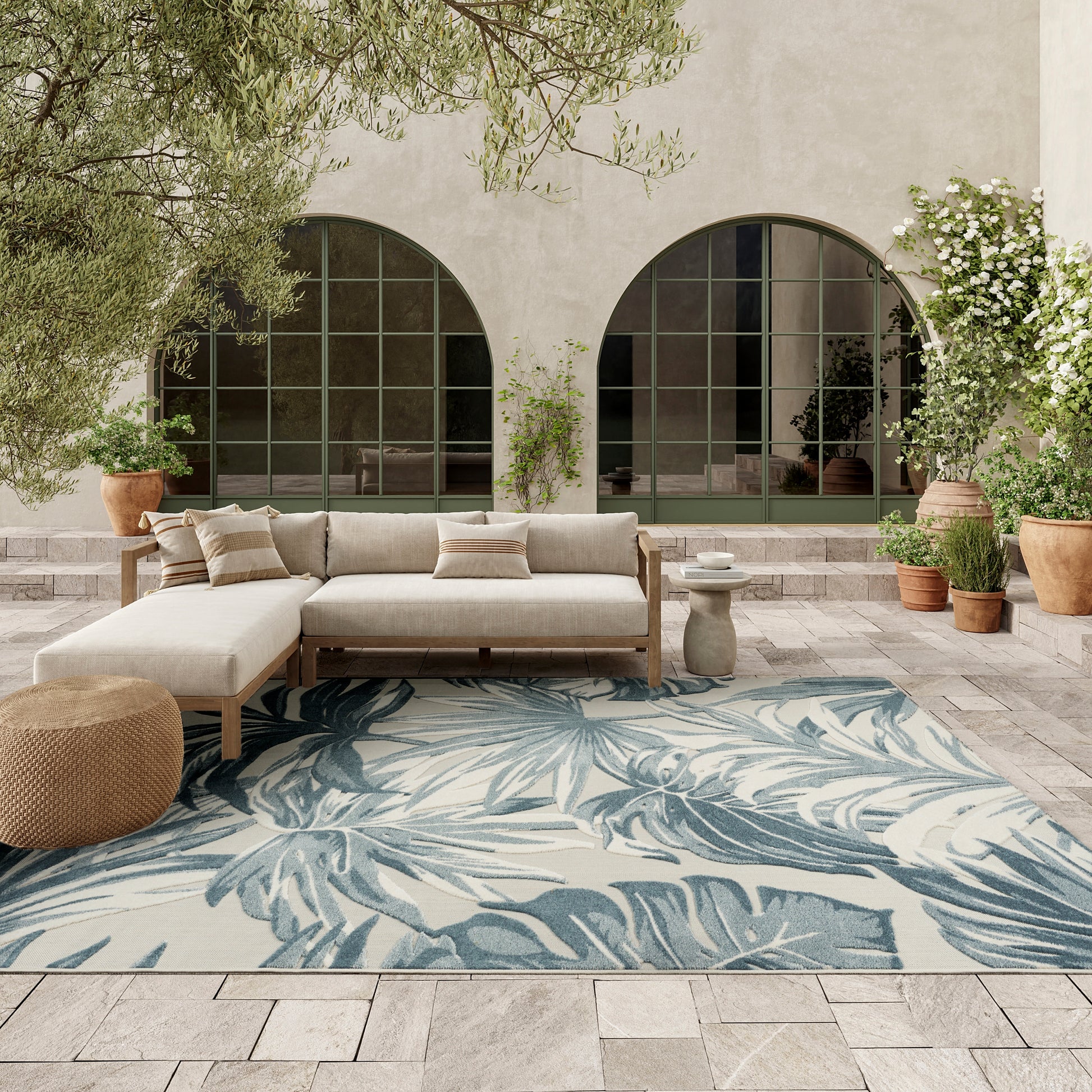 Outdoor beige sectional sofa with cushions on a blue palm-leaf rug, arched green-framed windows and potted plants.