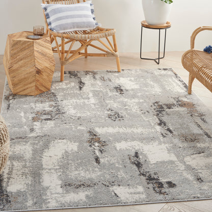 Gray & cream abstract area rug on light wood floor, with rattan chairs, striped pillow, and a plant stand.
