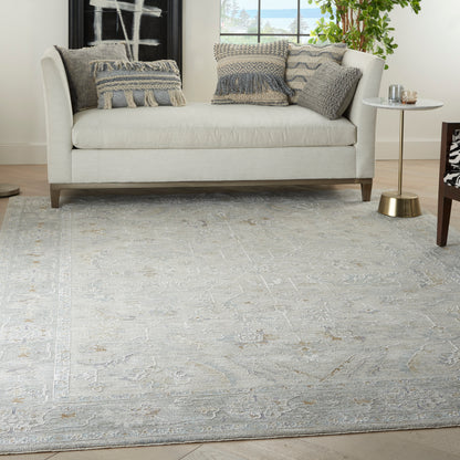 Light gray upholstered sofa with textured blue-gray and patterned pillows on a pale ornate rug; round gold side table.