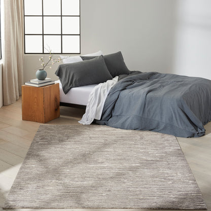 Modern bedroom with grey-blue duvet, dark-grey pillows, white sheets, wooden nightstand, vase, grid window, and beige rug.
