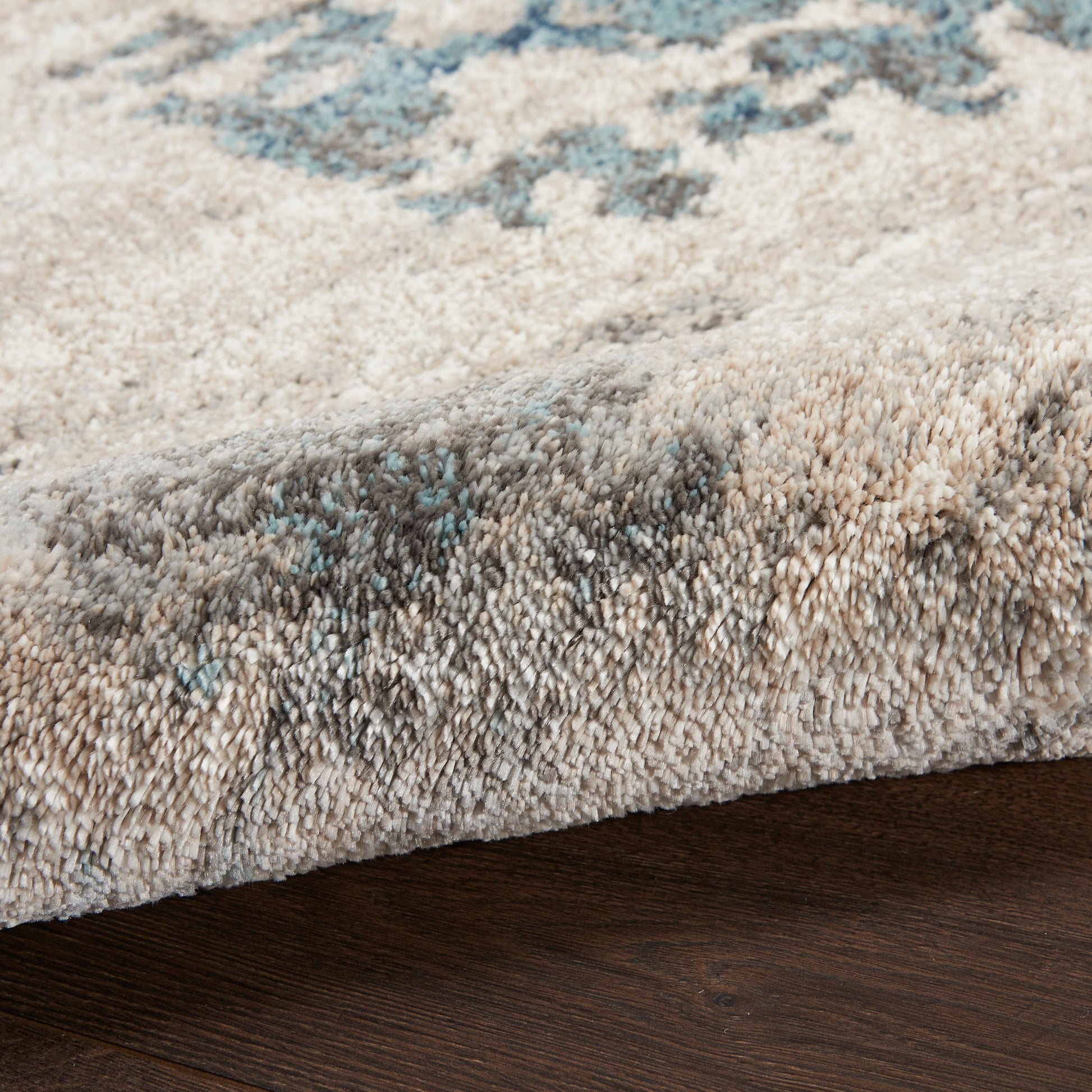 Plush beige-gray abstract area rug with blue accents, textured cut-pile & rolled edge on dark wood floor.