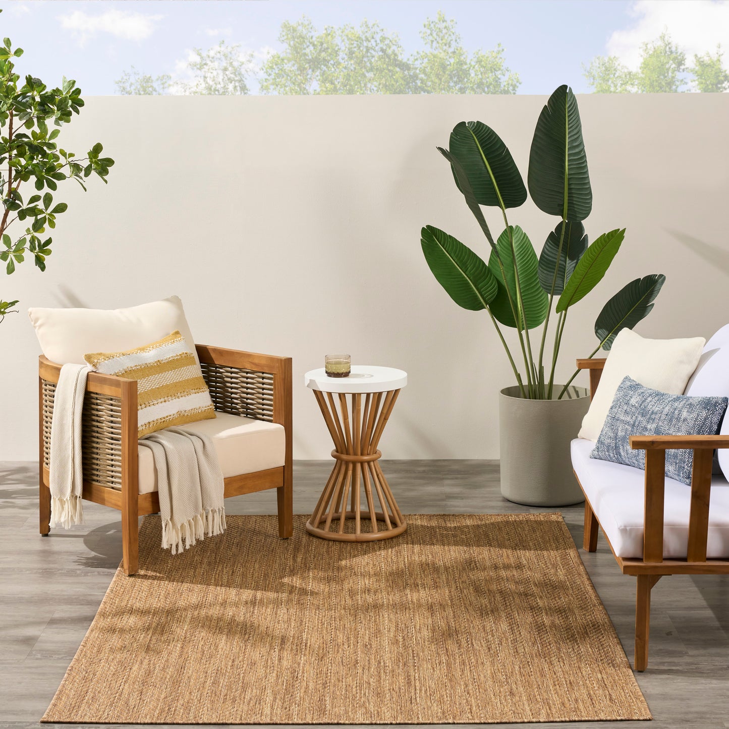5' x 7' jute rug in a living room scene with wooden armchairs, white cushions, striped pillow, beige throw, round white side table, and a large tropical plant.
