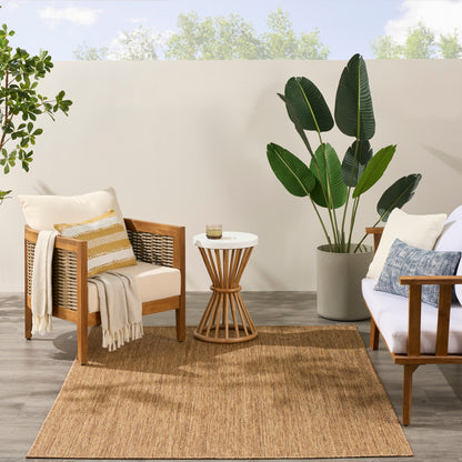 5' x 7' jute rug in a living room scene with wooden armchairs, white cushions, striped pillow, beige throw, round white side table, and a large tropical plant.