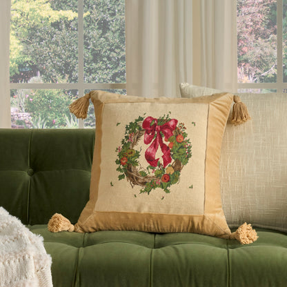 Golden-tan decorative pillow with Christmas wreath embroidery and a red bow, tassel fringes on a green tufted sofa.
