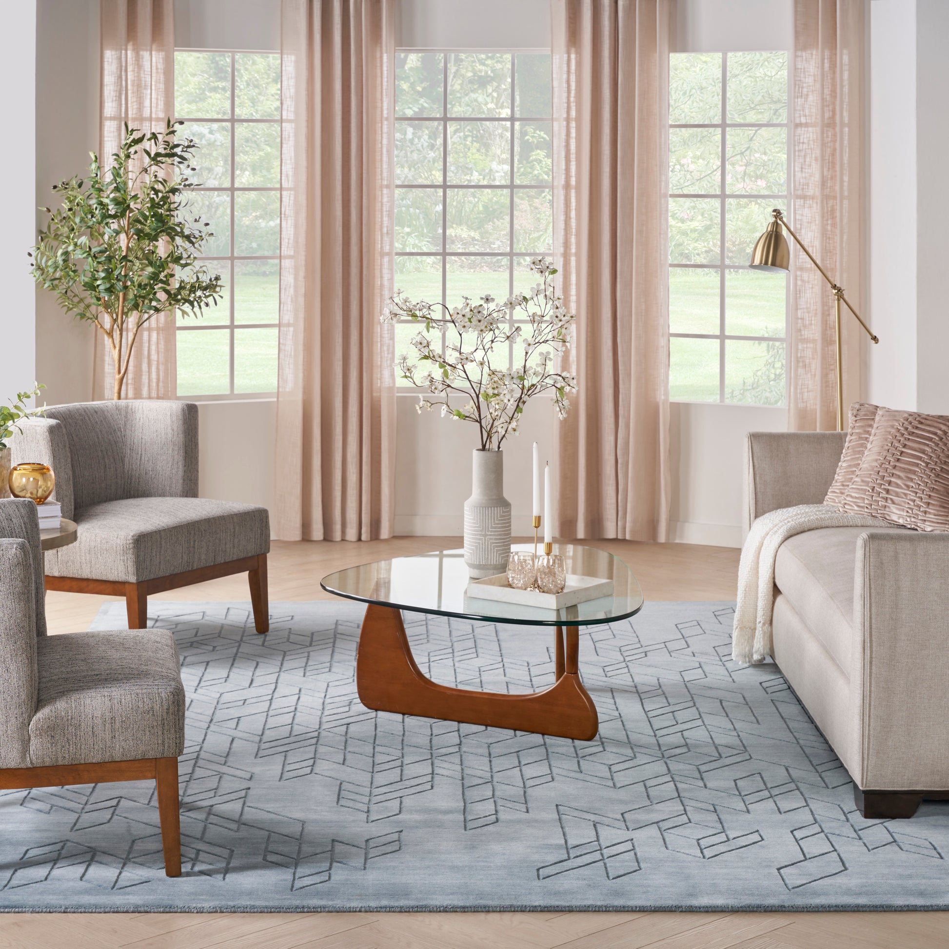 Cozy living room with beige curtains, glass coffee table on curved wood base, gray upholstered chairs, blue geometric rug.