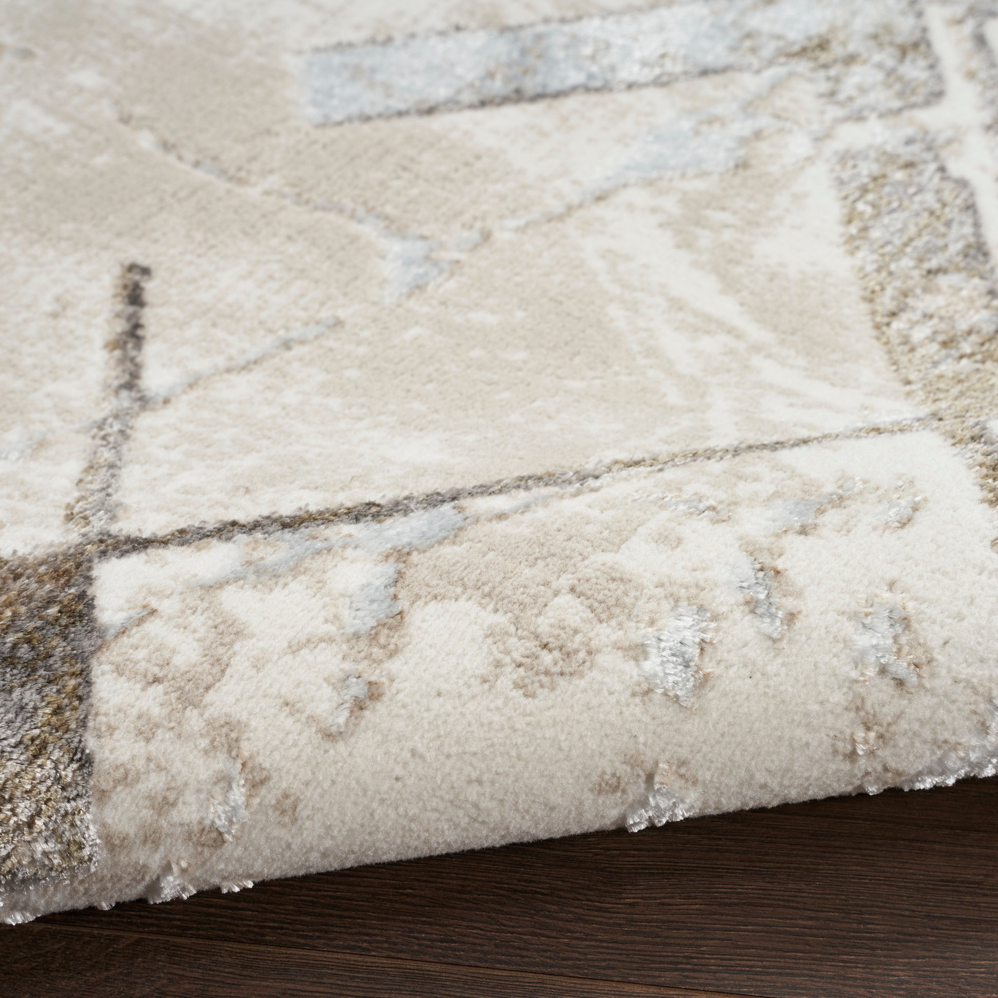 Beige abstract area rug with geometric patterns and light gray accents, folded corner on dark wood floor.