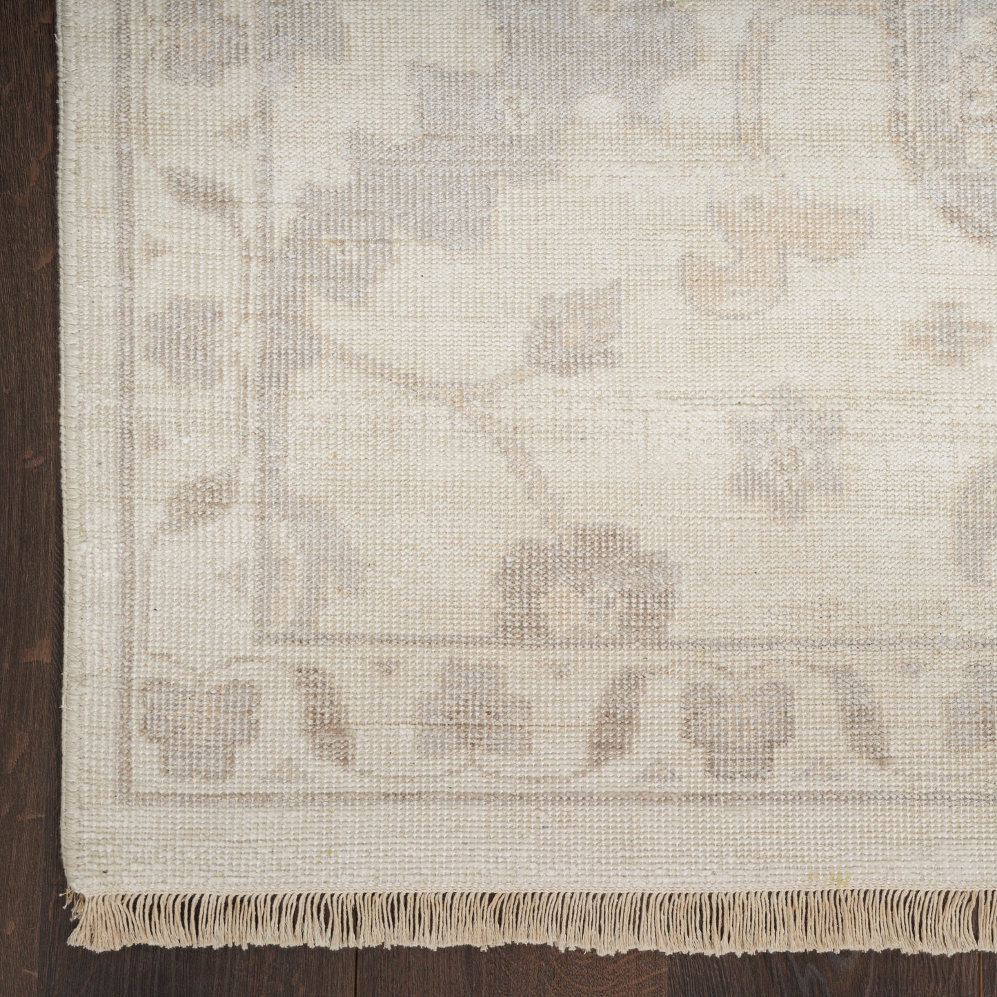 Beige woven rug with subtle floral border and fringe along bottom edge; dark wood floor on the left.