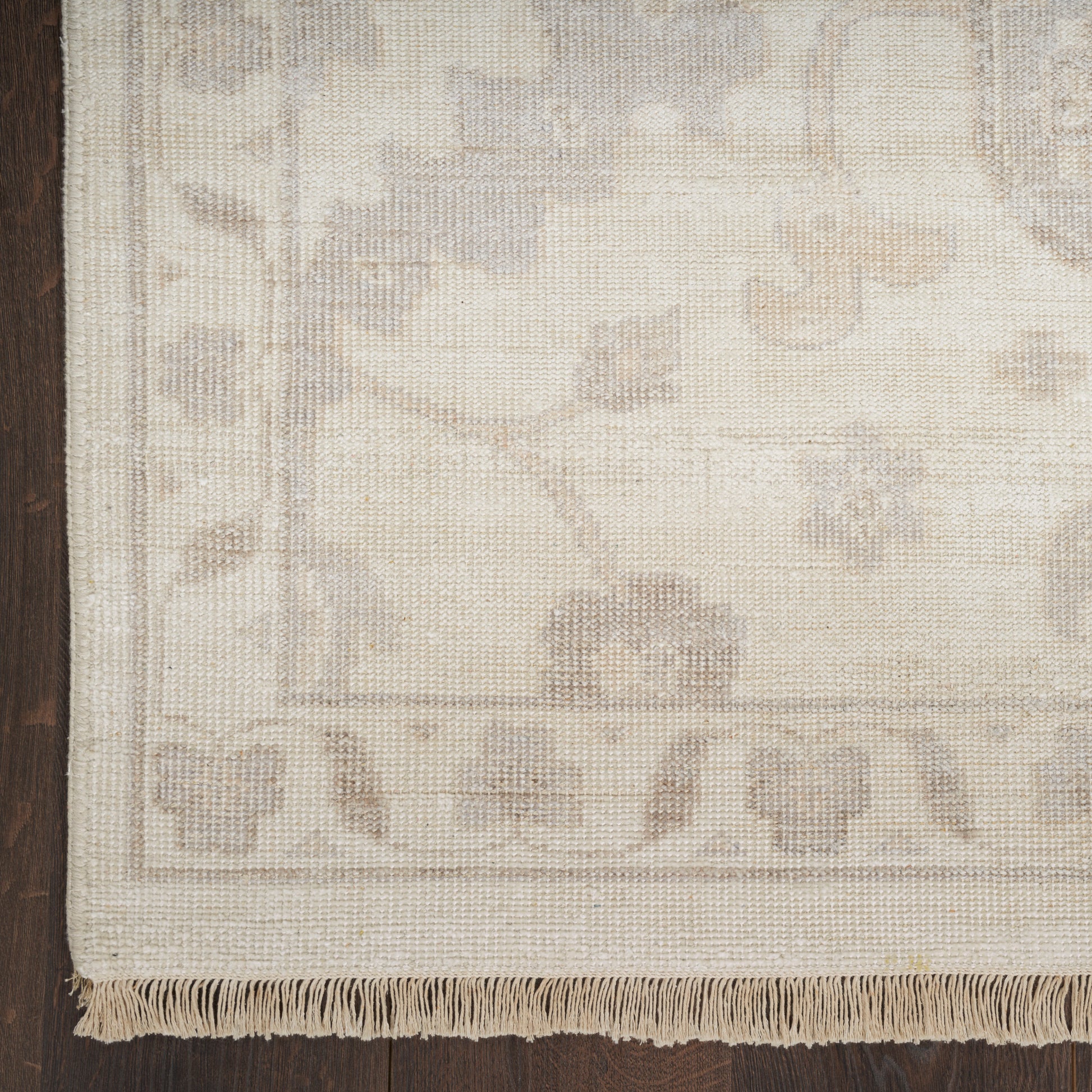 Beige woven rug with subtle floral border and fringe along bottom edge; dark wood floor on the left.