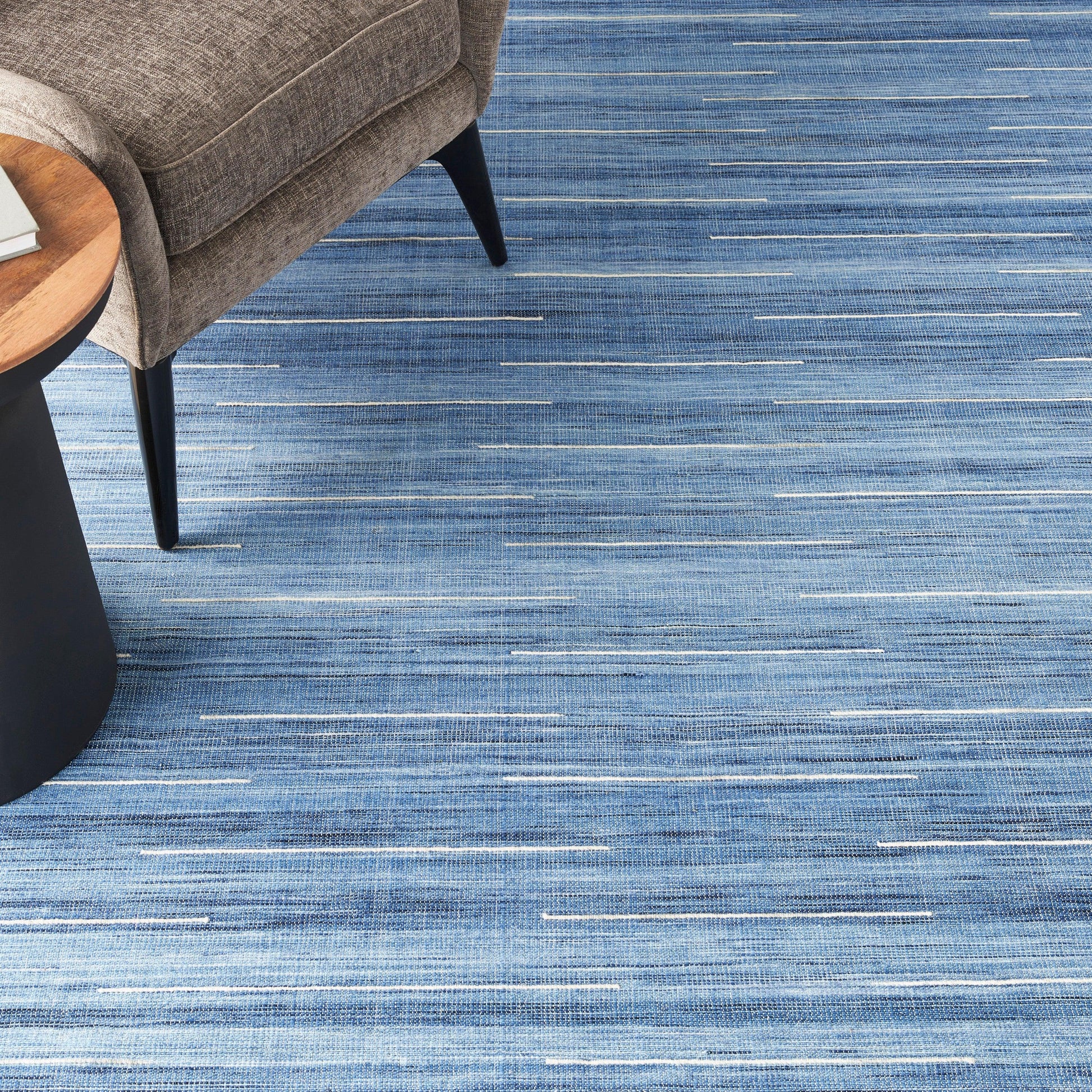 Blue striped area rug in varying light and dark blue tones under a beige upholstered chair with a round wooden table.