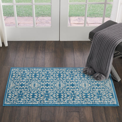 Rectangular teal-blue area rug with white damask floral pattern and blue border; draped dark gray knit throw on chair nearby.